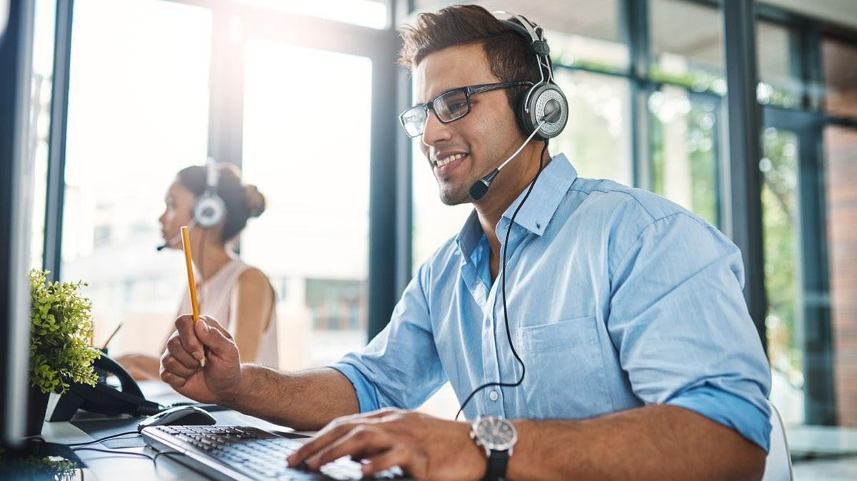 A smiling customer service representative wearing a headset and glasses, working at a computer.