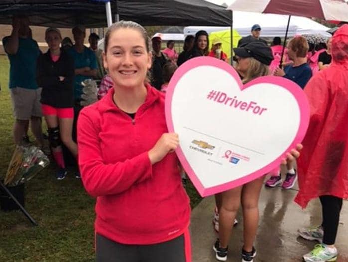Smiling young woman holding an #IDriveFor heart sign at a pink event.