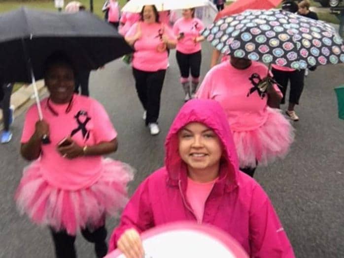 Women wearing pink shirts and tutus walk in the rain with umbrellas.