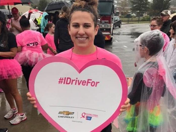 Smiling woman in pink holds a large, blank #IDriveFor heart sign.