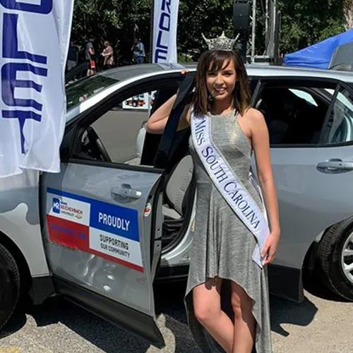 A woman in a pageant sash stands beside a grey SUV with event decals