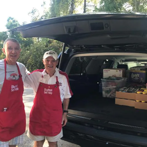 Two people in red aprons stand by the open trunk of a van full of supplies