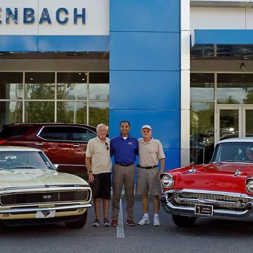 Three men stand between a yellow Camaro and red classic car at a dealership