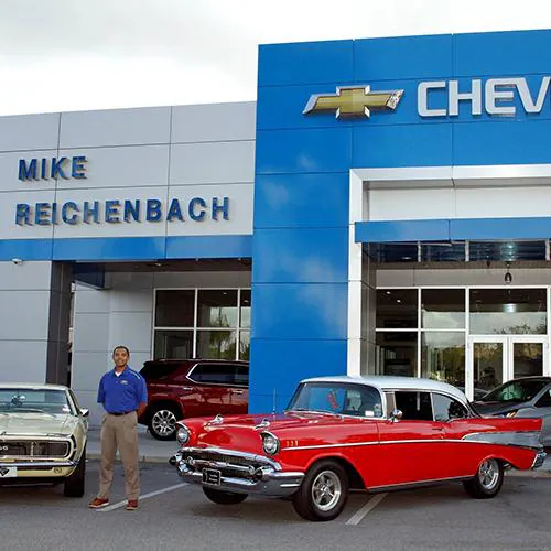A man stands next to a red and white classic Chevrolet outside Mike Reichenbach Chevrolet.