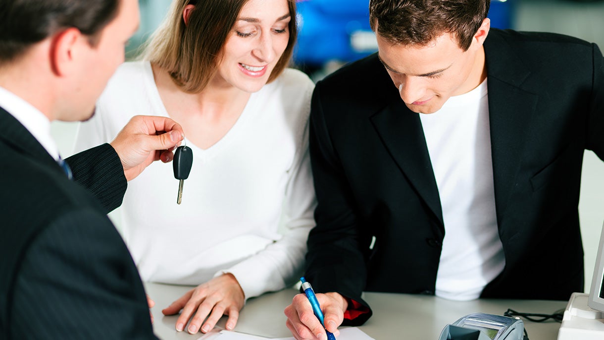 A couple is signing papers while a salesman hands over car keys.